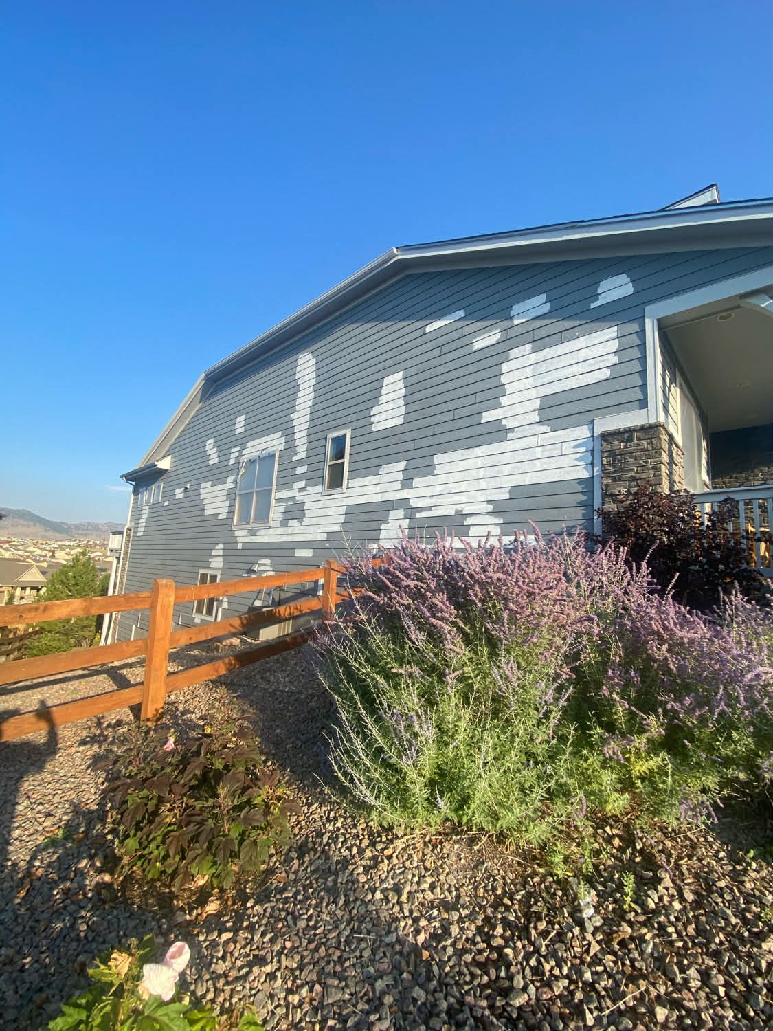 A house with faded, peeling blue siding under a bright blue sky, with a wooden fence and landscaping in the foreground.