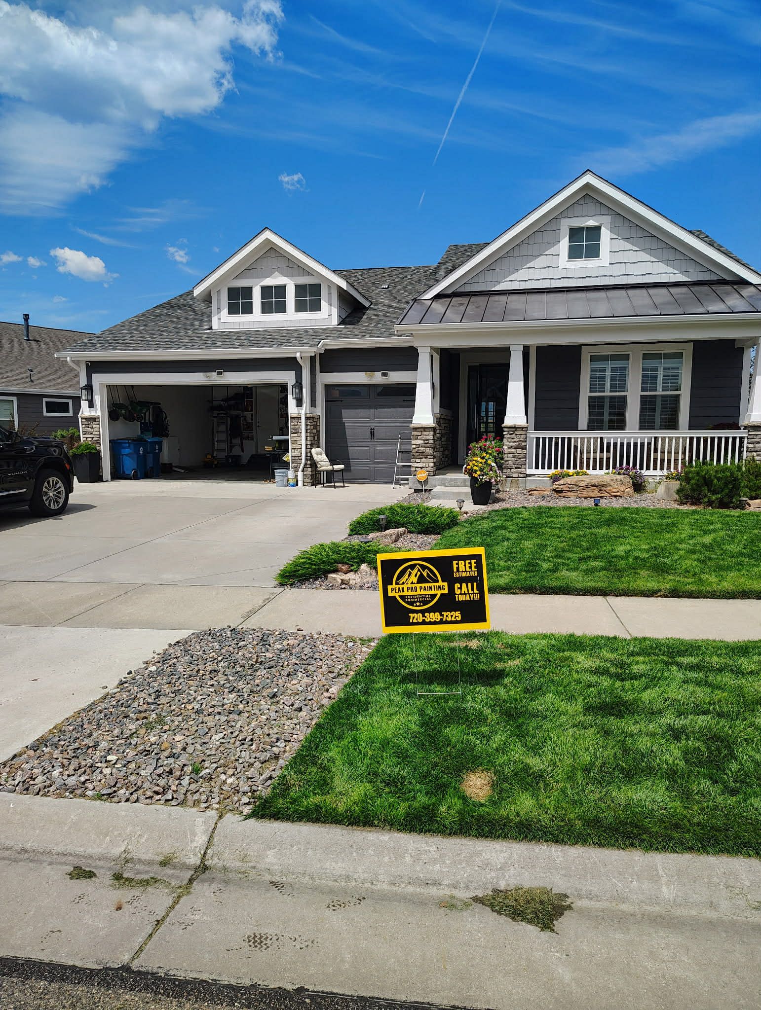 A grey, two-story suburban house with a garage and a white front porch under a bright, partly cloudy blue sky.