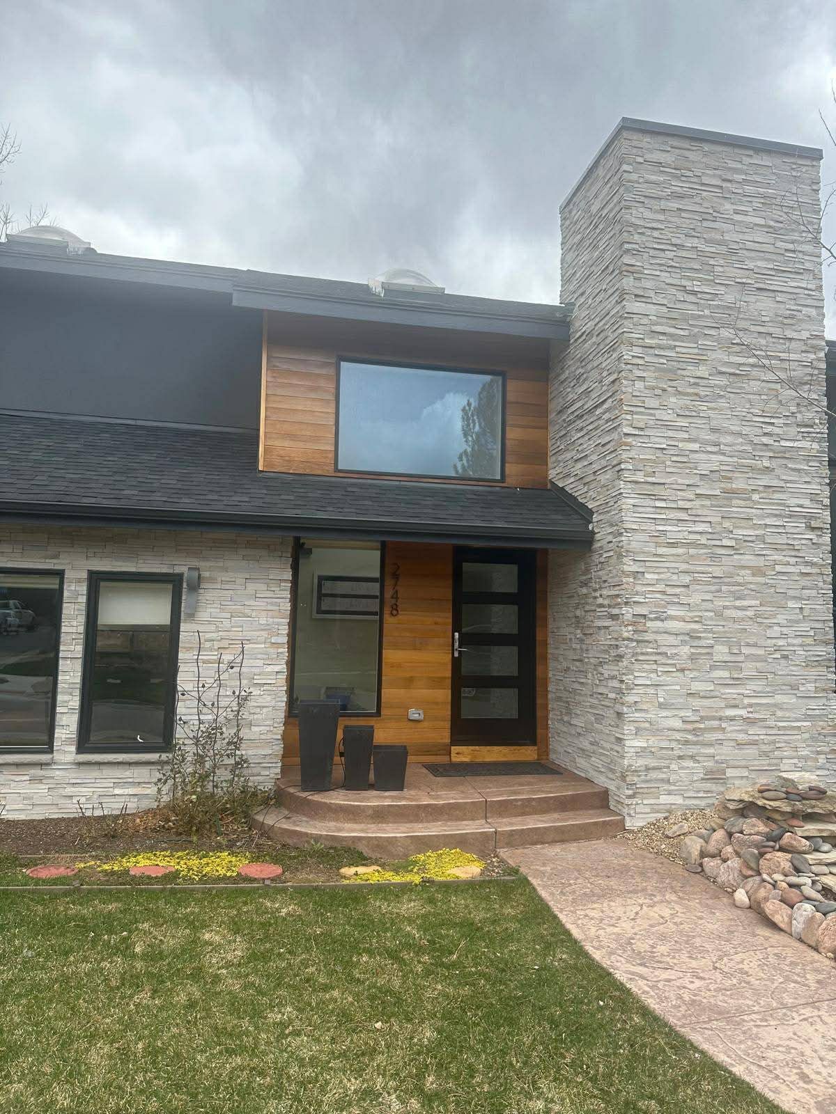 A modern home entrance with a white stone chimney, wood-paneled siding, a dark front door, and a paved walkway.