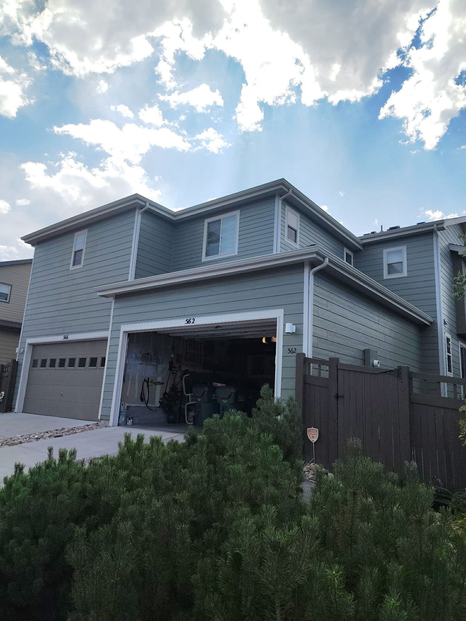 A two-story grey house with a visible garage, white trim, and a bush in the foreground under a cloudy blue sky.