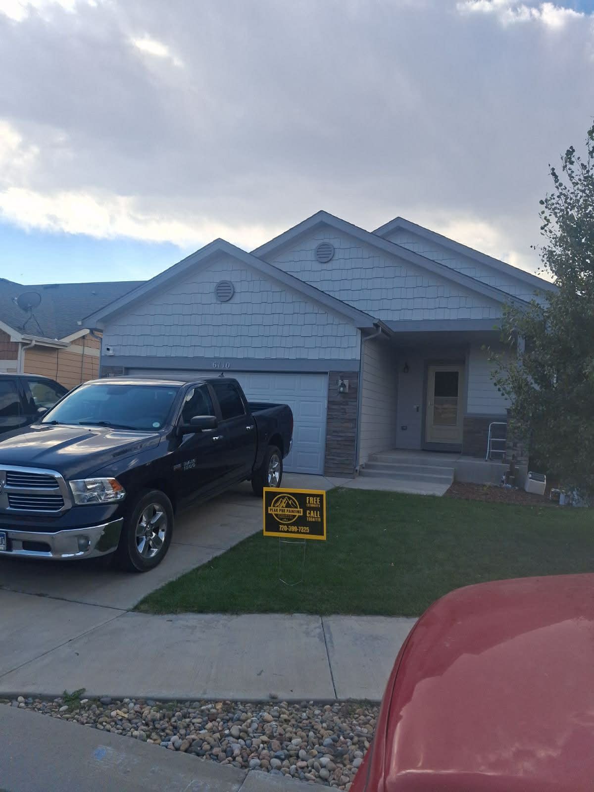 A black pickup truck parked in the driveway of a light blue, one-story suburban home with a yard sign on the grass.