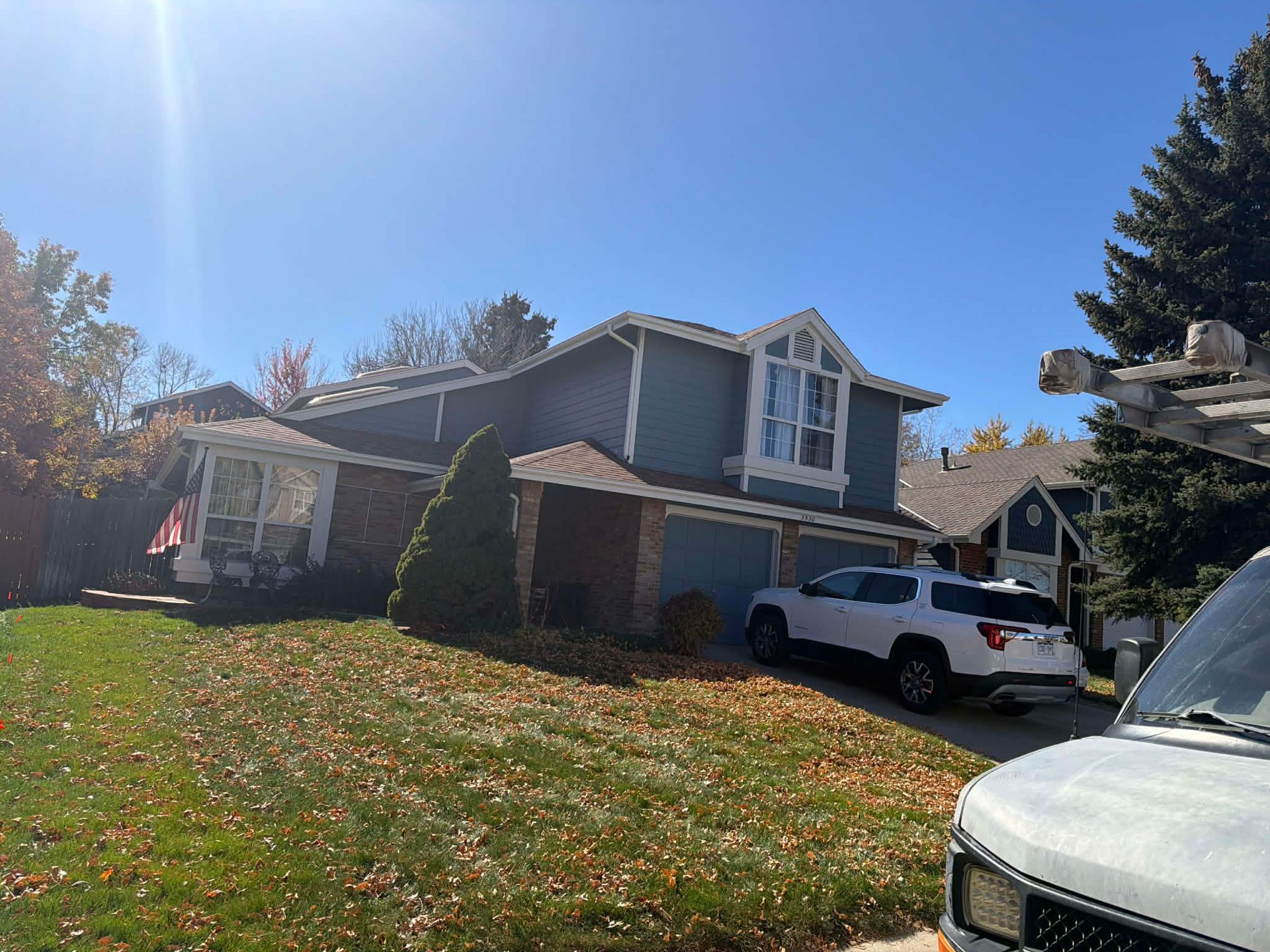 A two-story blue house with a brick accent, a white SUV in the driveway, and a lawn covered in autumn leaves.