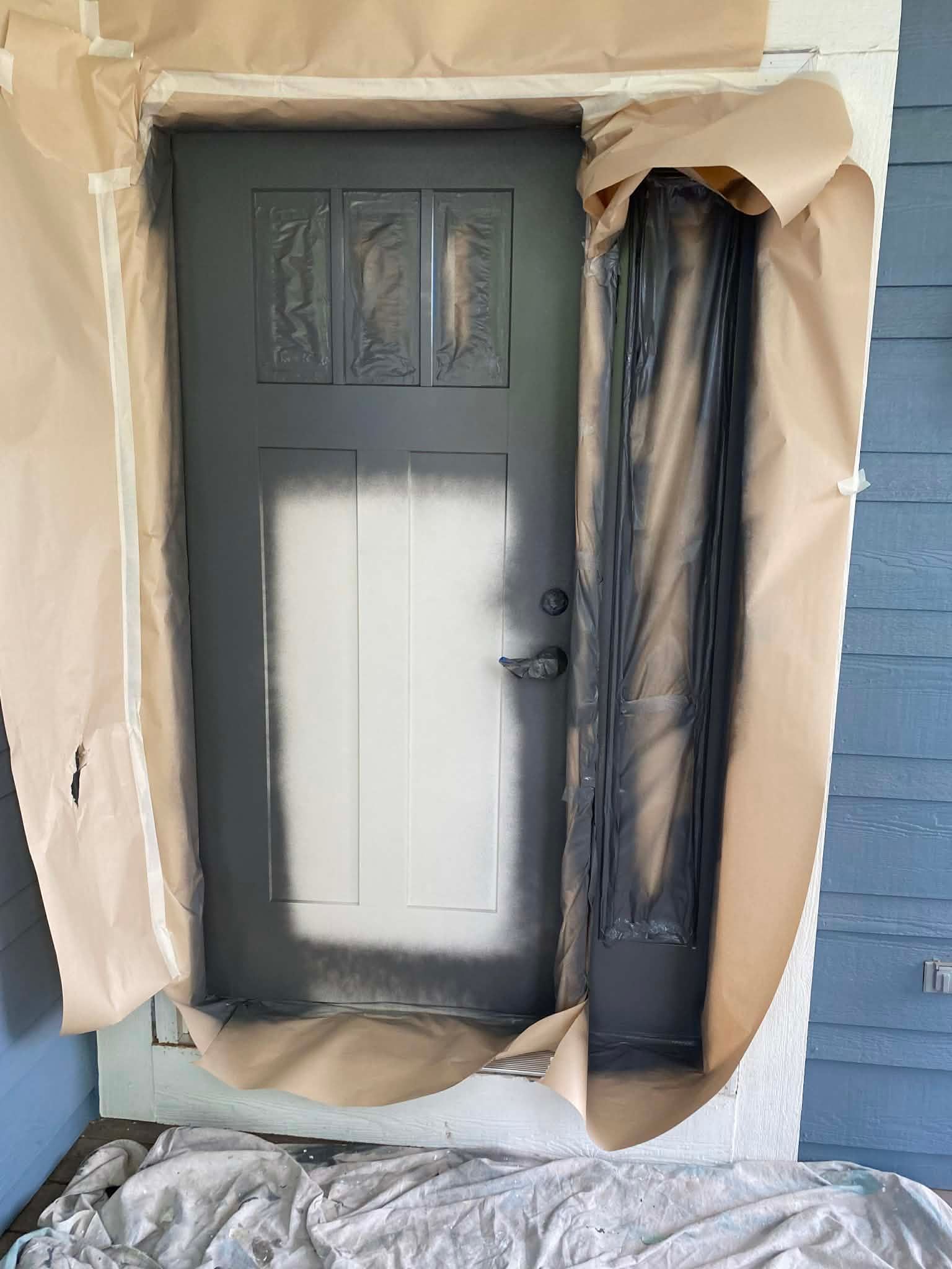 A dark grey front door being painted, with masking paper around the frame and a white primer patch in the center.