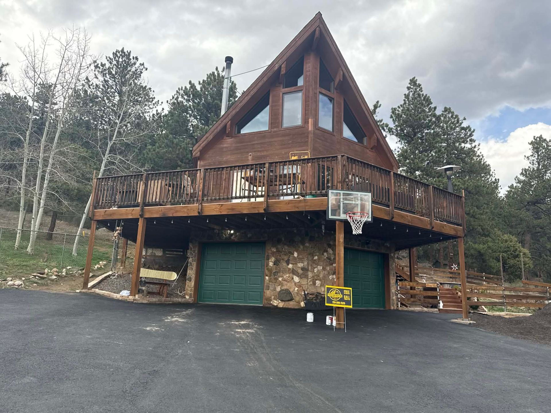 A wooden A-frame house built above a two-car stone garage, featuring a large deck and surrounded by pine trees.