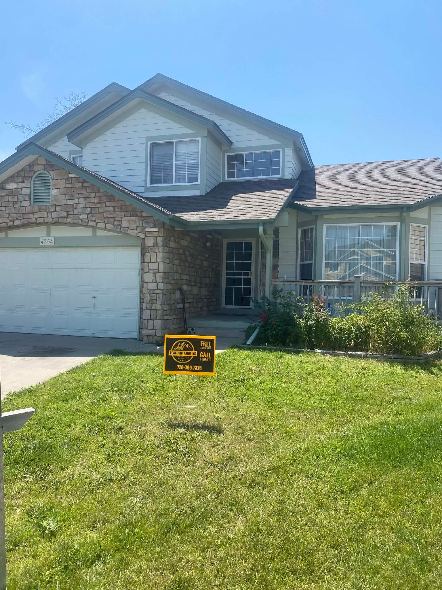A two-story suburban home with a stone-accented facade, attached garage, and a lawn sign under a clear blue sky.