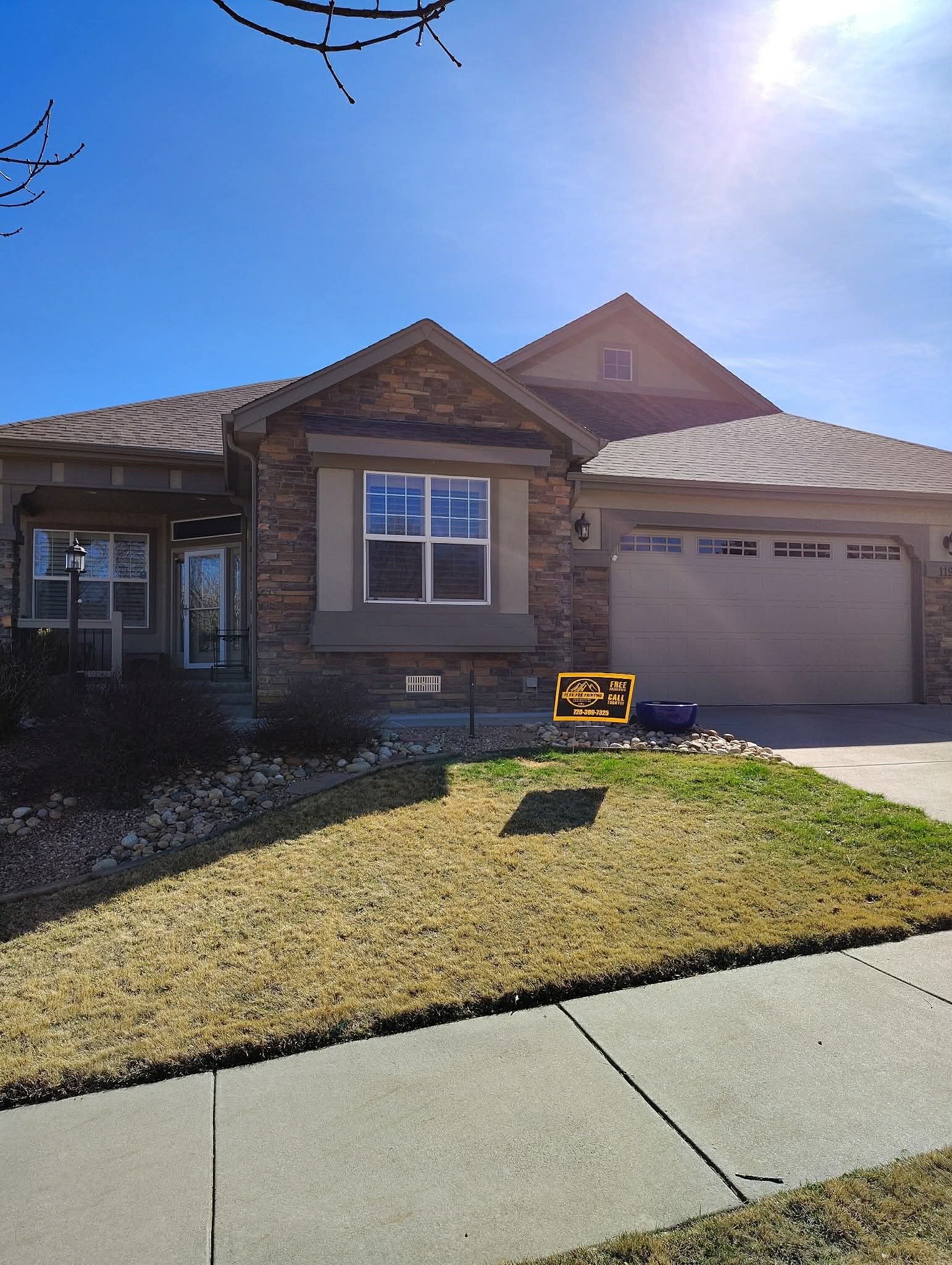 A tan, single-story stone and stucco house under a bright blue sky with a concrete driveway and small front lawn.