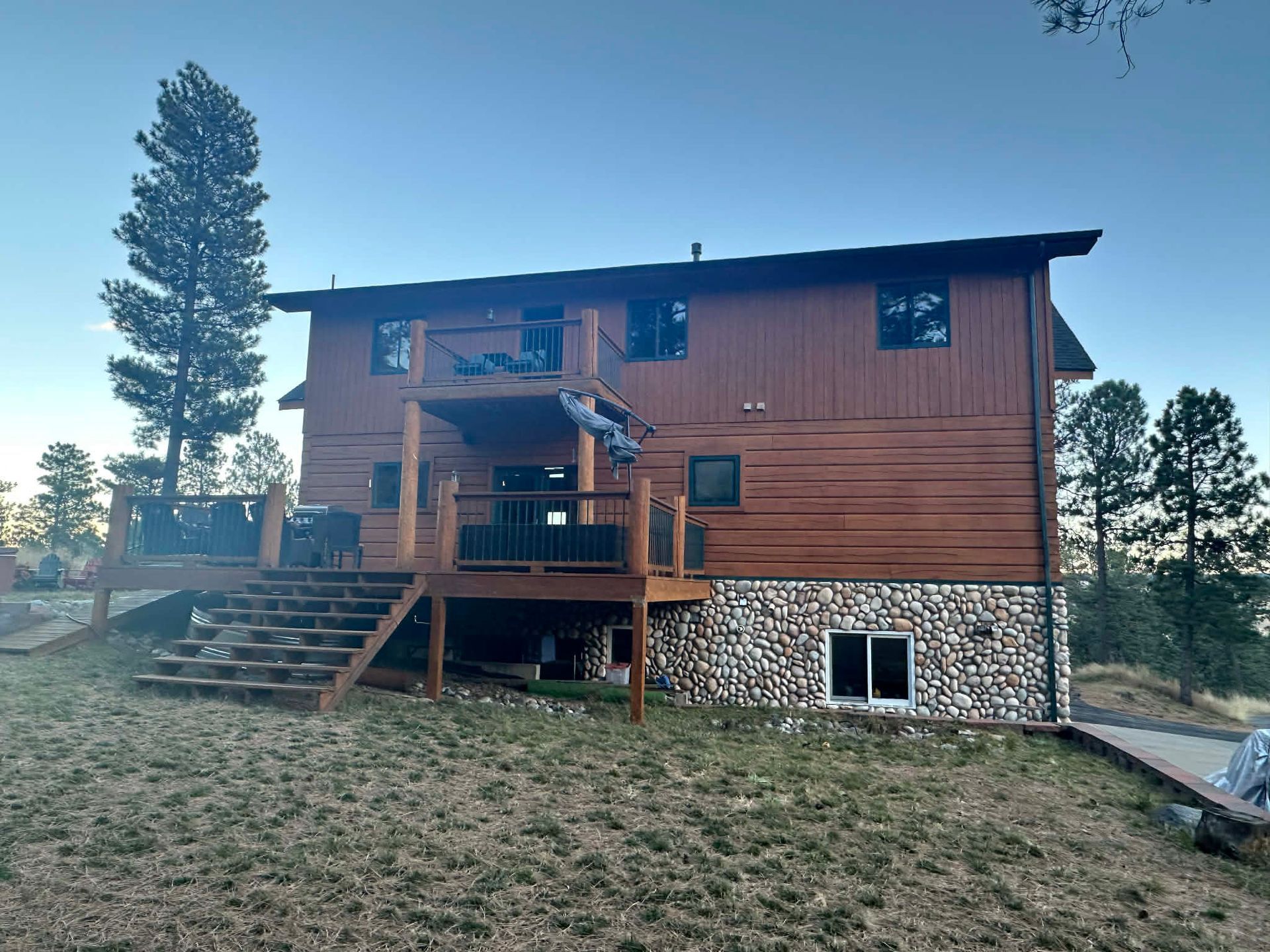 Two-story wood cabin with a stone foundation, a raised deck, stairs, and surrounding trees under a clear blue sky.