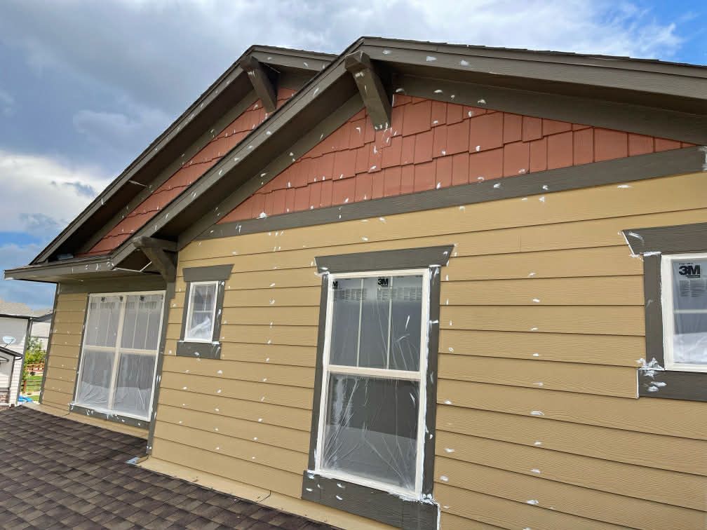 A partially constructed house exterior with tan horizontal siding, reddish-brown shingles in the gable, and dark trim.