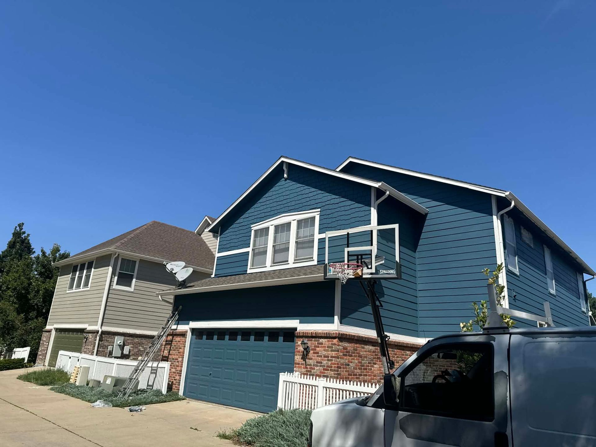 Two houses with blue and tan siding stand on a suburban street under a clear blue sky, with a van and basketball hoop.