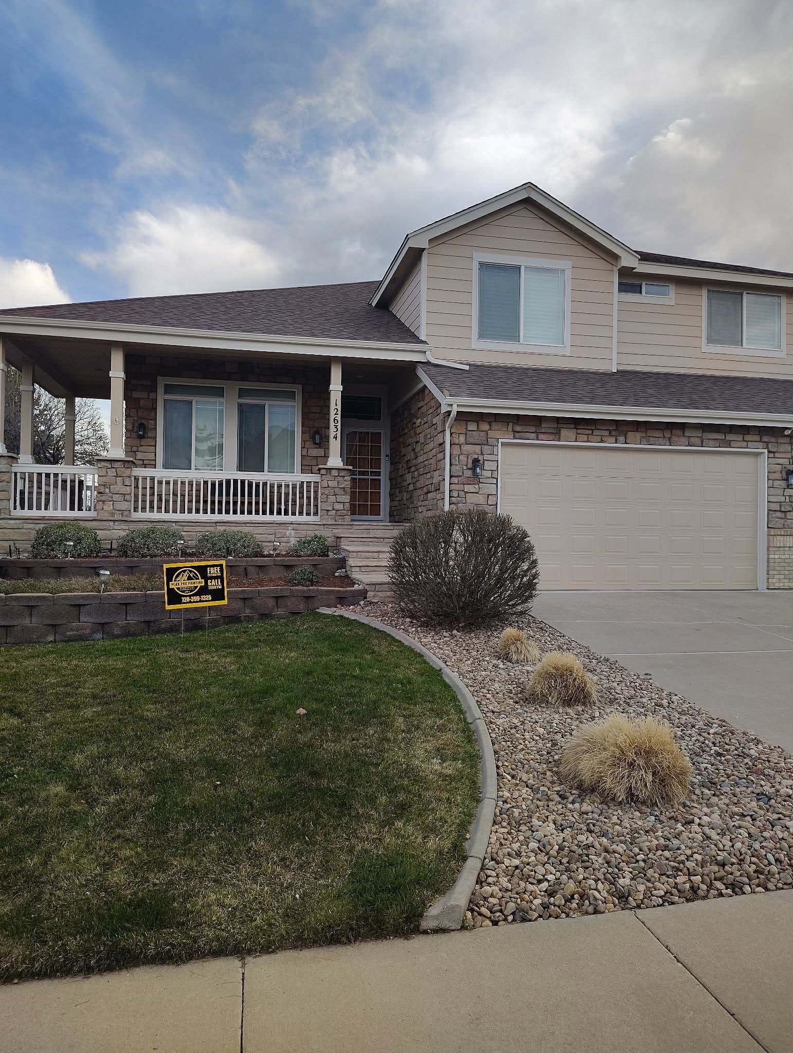 A split-level home with tan siding, a brown shingle roof, a two-car garage, and a front porch, featuring a gravel yard.