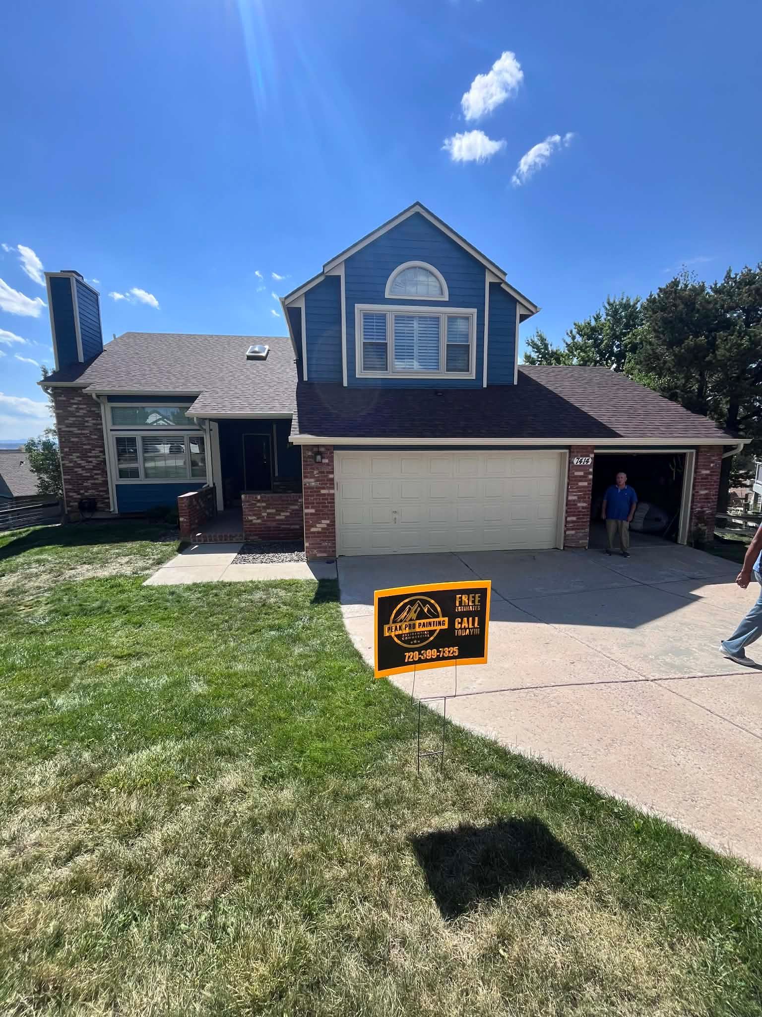A two-story blue house with a brick base and a garage, featuring a lawn sign in front under a clear blue sky.