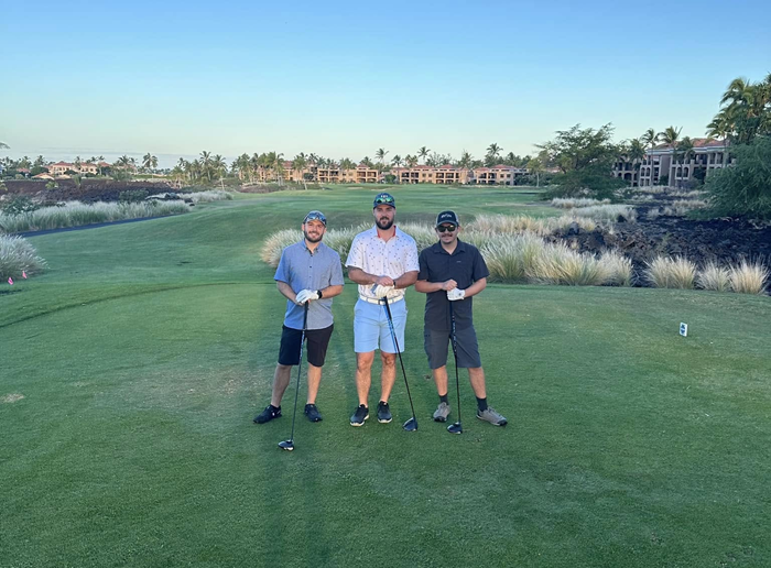 Three people stand on a sunny golf course, each holding a golf club, with buildings and native grasses in the background.