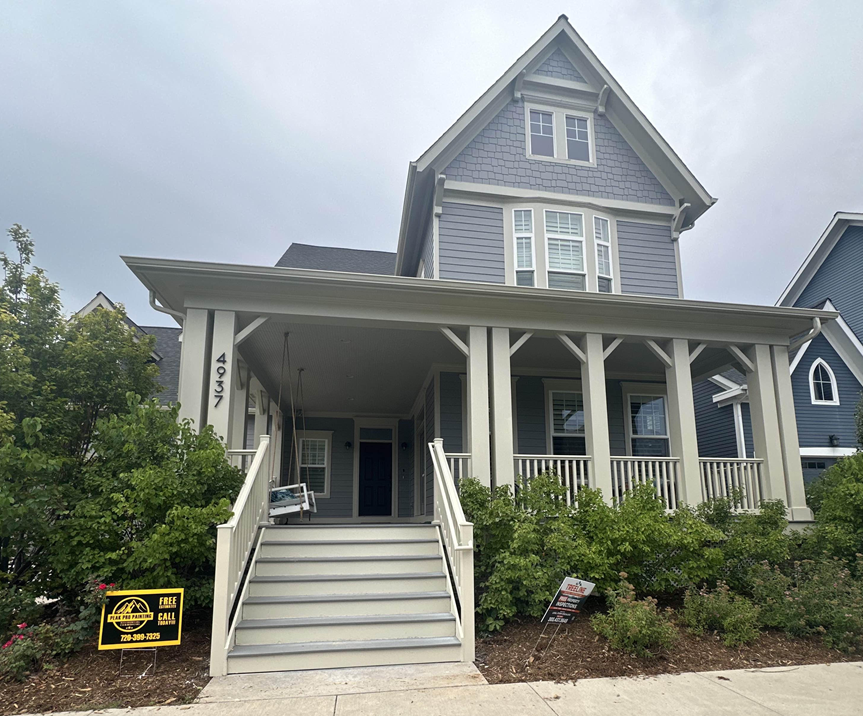 A blue two-story house with a white wraparound porch, front steps, and a yard with green bushes under a cloudy sky.