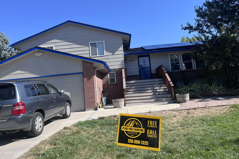 A two-story grey suburban house with blue trim, a grey SUV in the driveway, and a yellow roofing company yard sign.