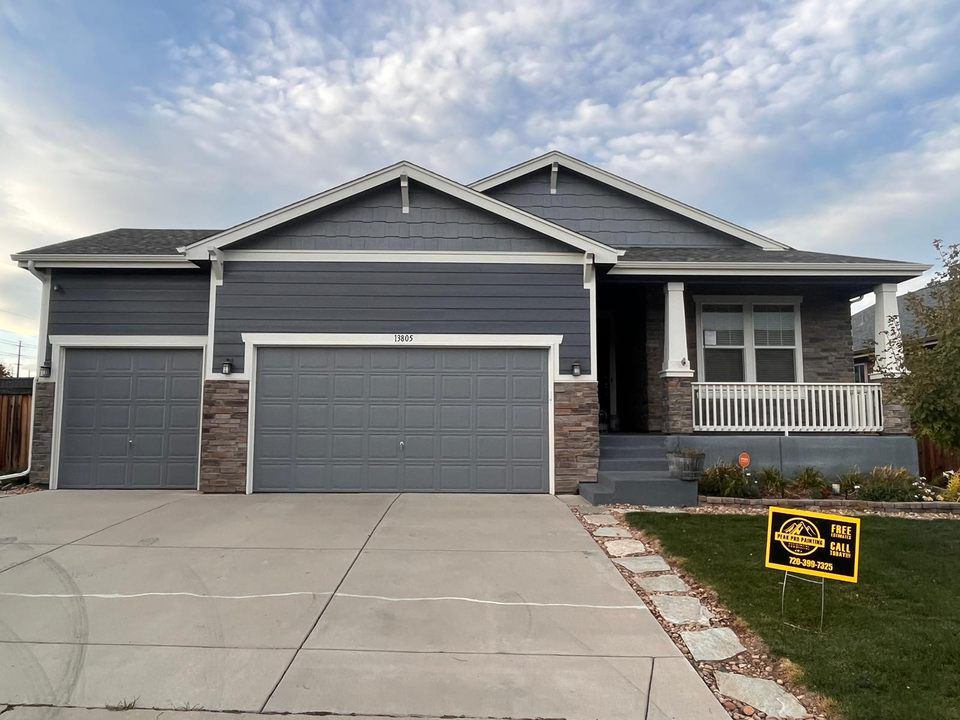 A modern, gray-sided suburban home with a three-car garage, stone accents, and a front porch, viewed from the driveway.