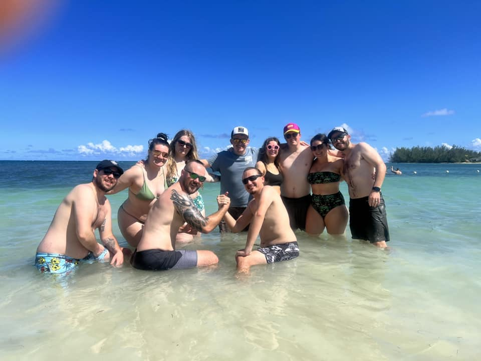 A group of eleven people posing together in shallow, clear turquoise ocean water on a sunny day.