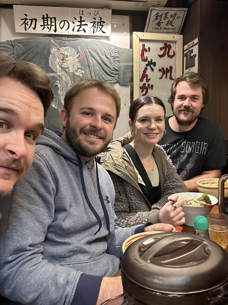 A group of four smiling people sitting at a table in a restaurant with Japanese signage on the wall behind them.