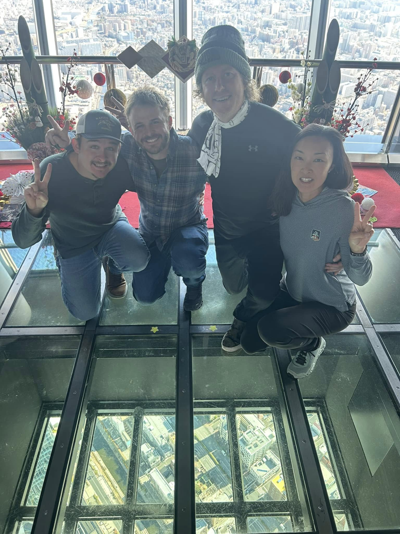 Four people pose on a glass floor with an aerial view of a city below, smiling and making peace signs.