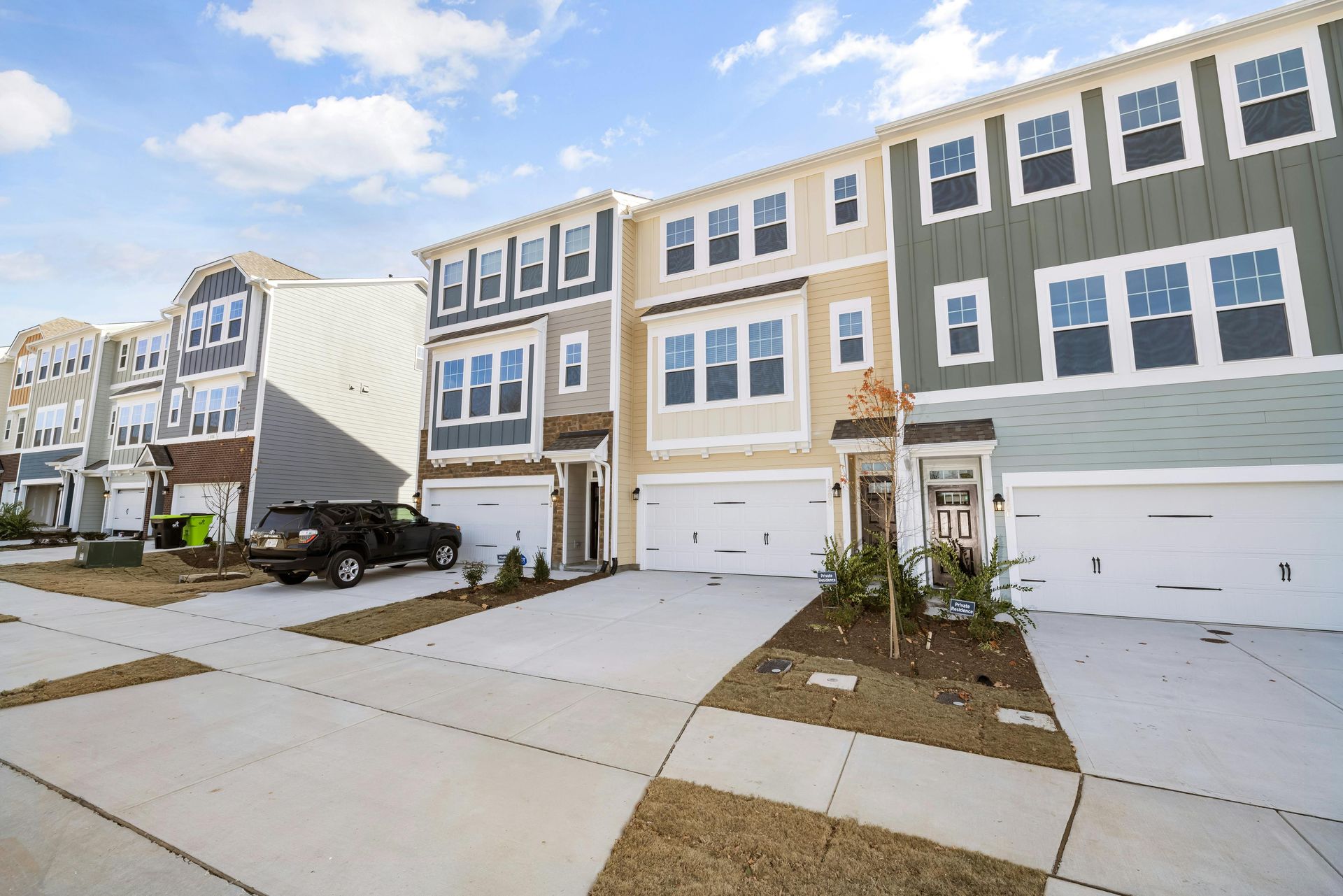 A row of modern three-story townhouses with beige, grey, and cream siding, white garage doors, and a parked black SUV.