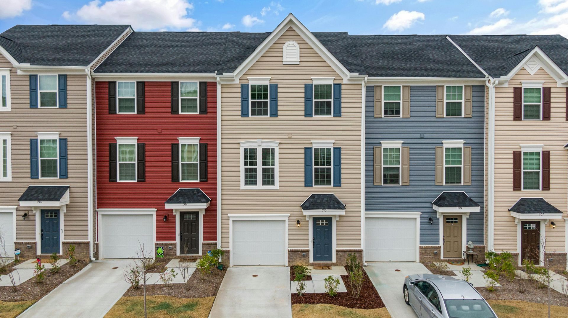 A row of colorful townhouses with white garage doors and paved driveways under a cloudy blue sky.