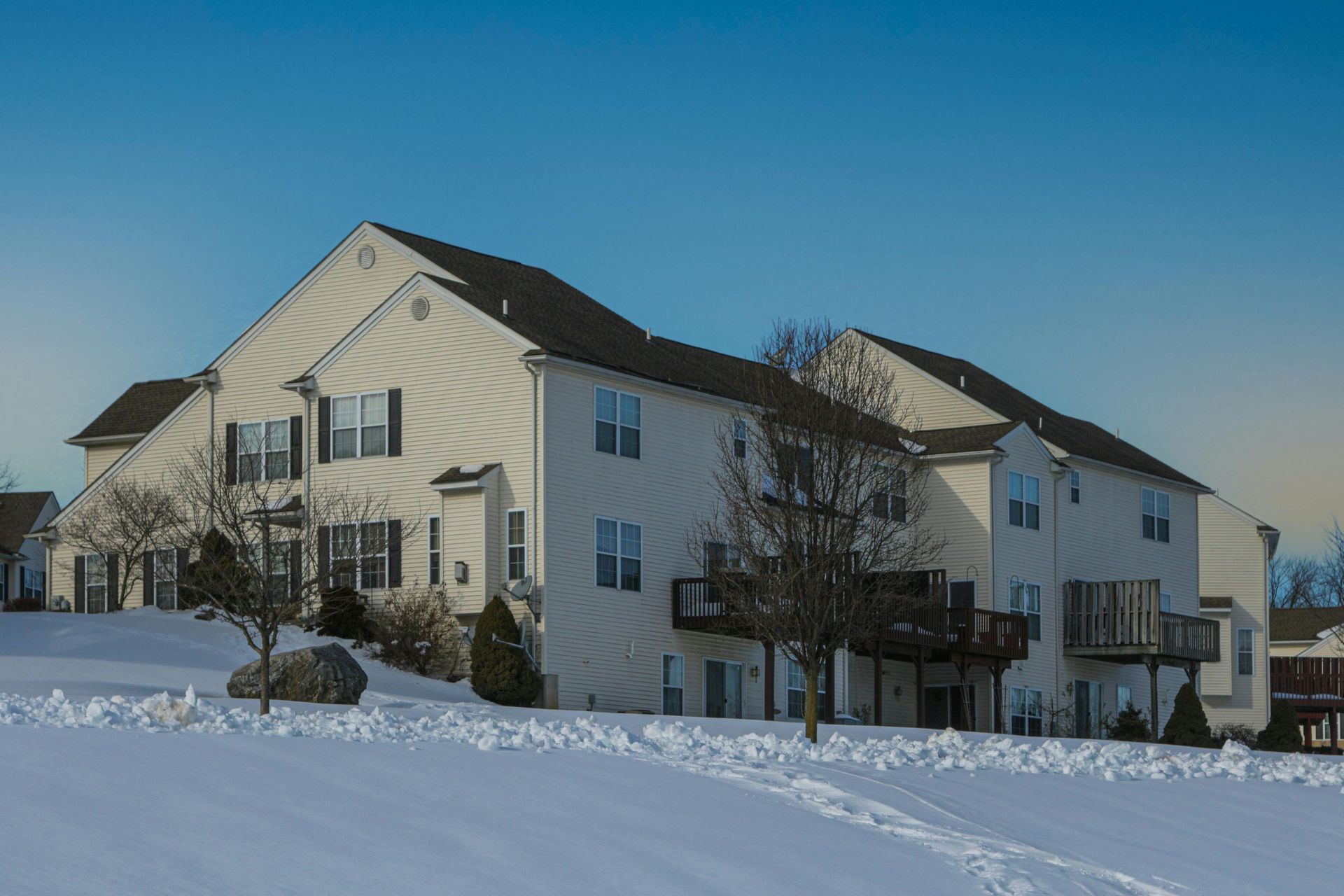 A multi-story apartment building with beige siding and dark roofs set against a snowy, sunlit landscape.