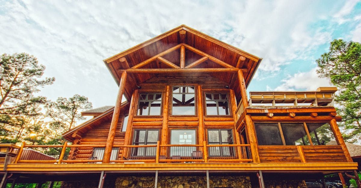 A large, multi-story wooden log cabin house with a wrap-around deck, multiple windows, and a forest setting.