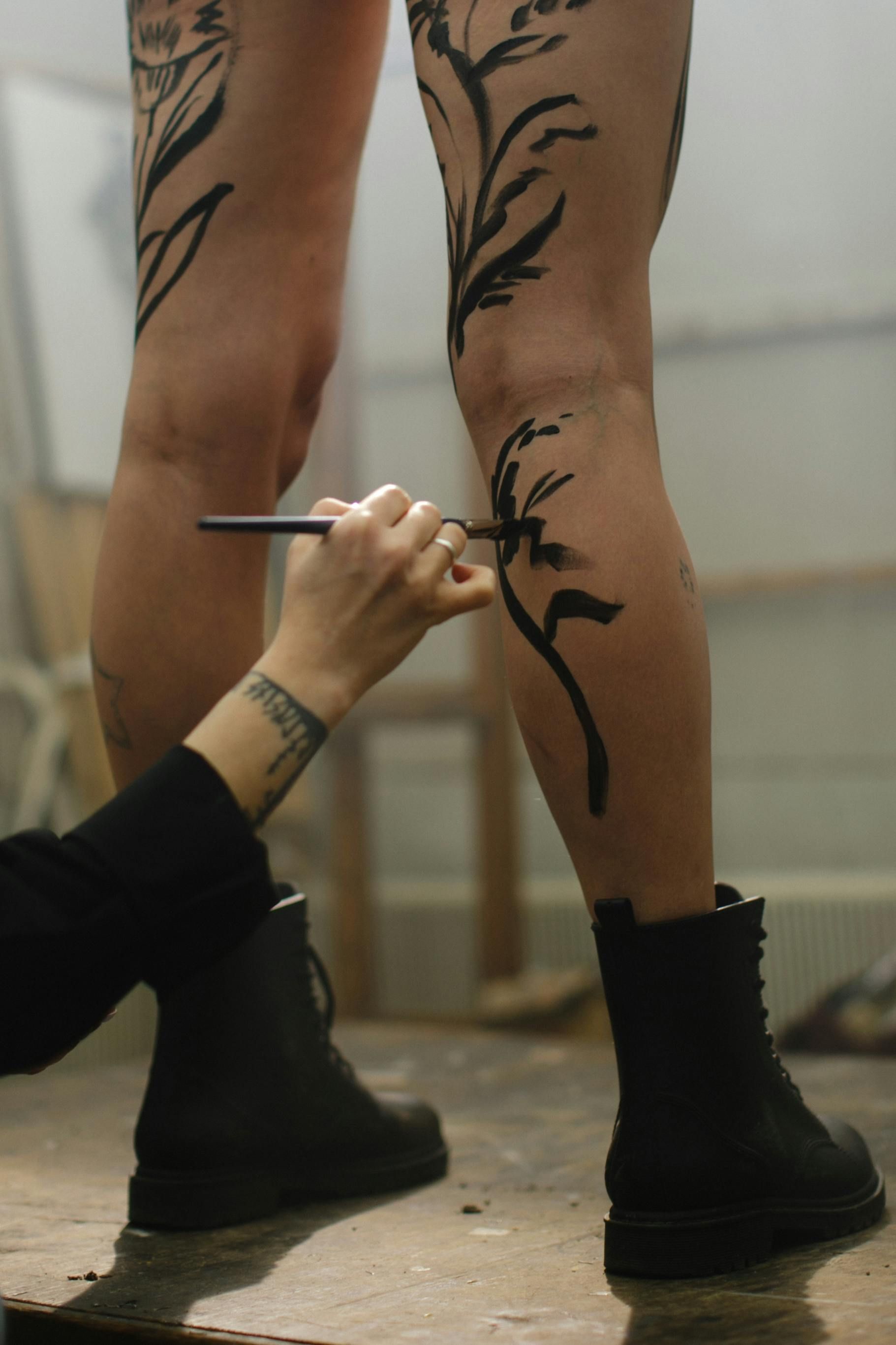 A hand uses a brush to paint a black, floral design onto a person's leg in an art studio setting.