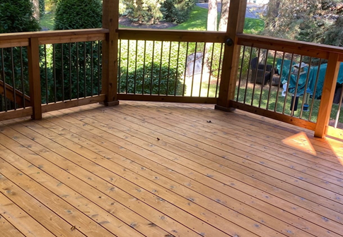 A wooden deck with a railed perimeter overlooks a tree-filled backyard in a sunny, outdoor setting.
