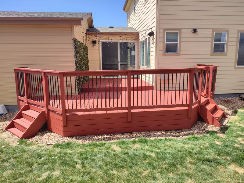 A raised, hexagonal wooden deck painted reddish-brown with railings and stairs, attached to a two-story beige house.