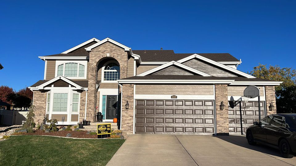 A two-story suburban home with tan siding, stone accents, a multi-car garage, and a driveway under a clear blue sky.