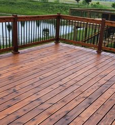 A freshly stained wooden deck with black metal railings overlooking a pond and green lawn.