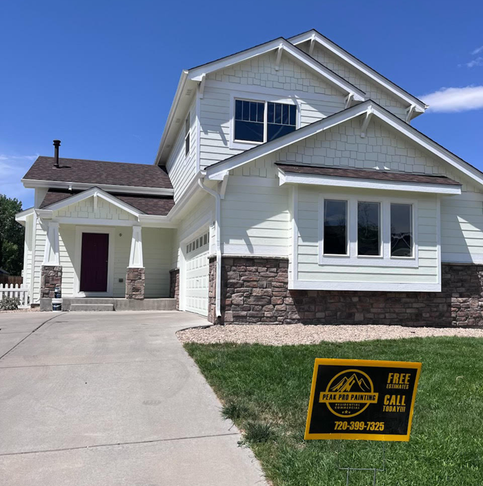 A two-story light-colored house with stone accents, a concrete driveway, and a painting service sign on the front lawn.
