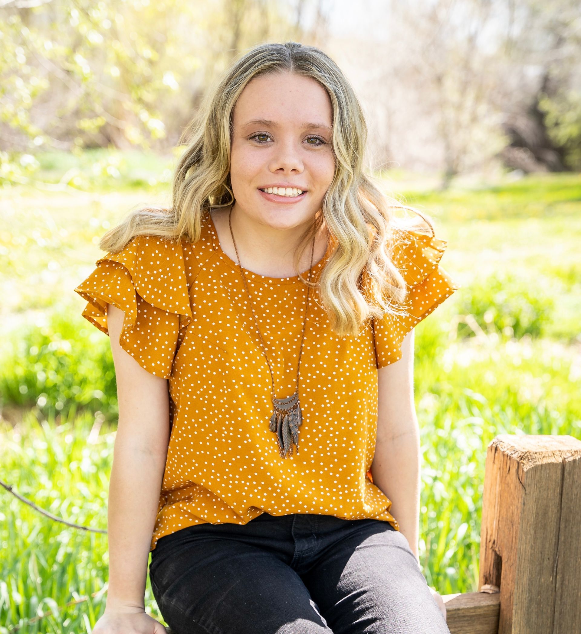 Woman with blond hair in a yellow polka-dot top, smiling, sitting on a wooden fence outdoors.