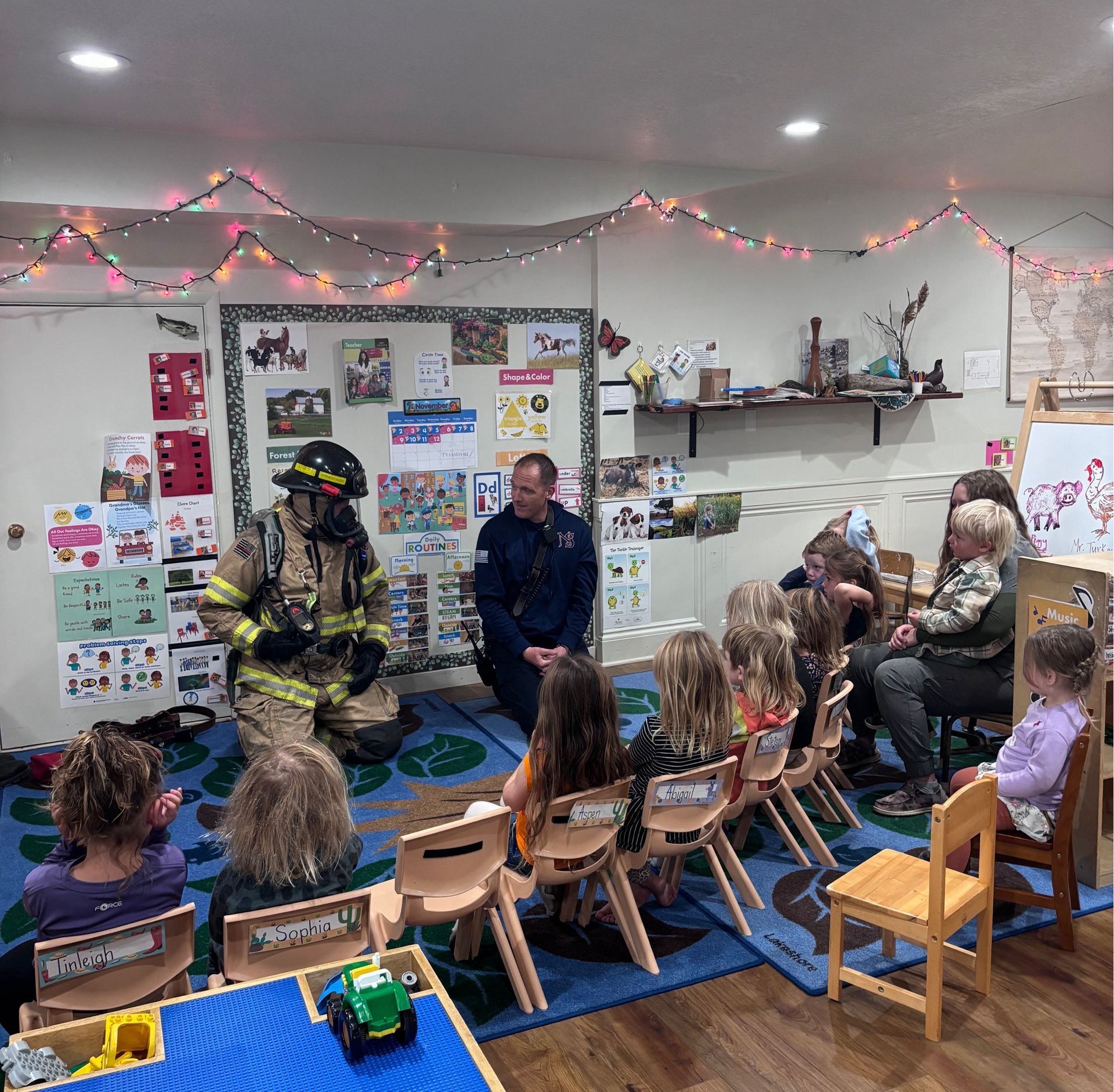 Firefighters with children in a classroom; one in gear, one seated; small chairs and decorations.