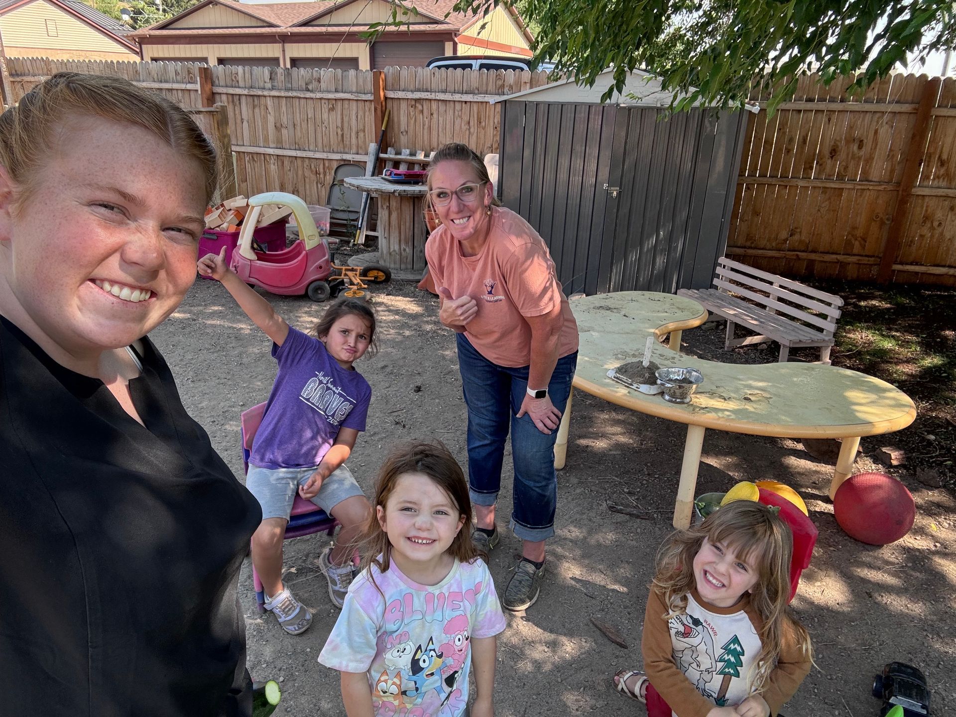 Group of people smiling in a fenced outdoor area; includes children, adults, and toys.
