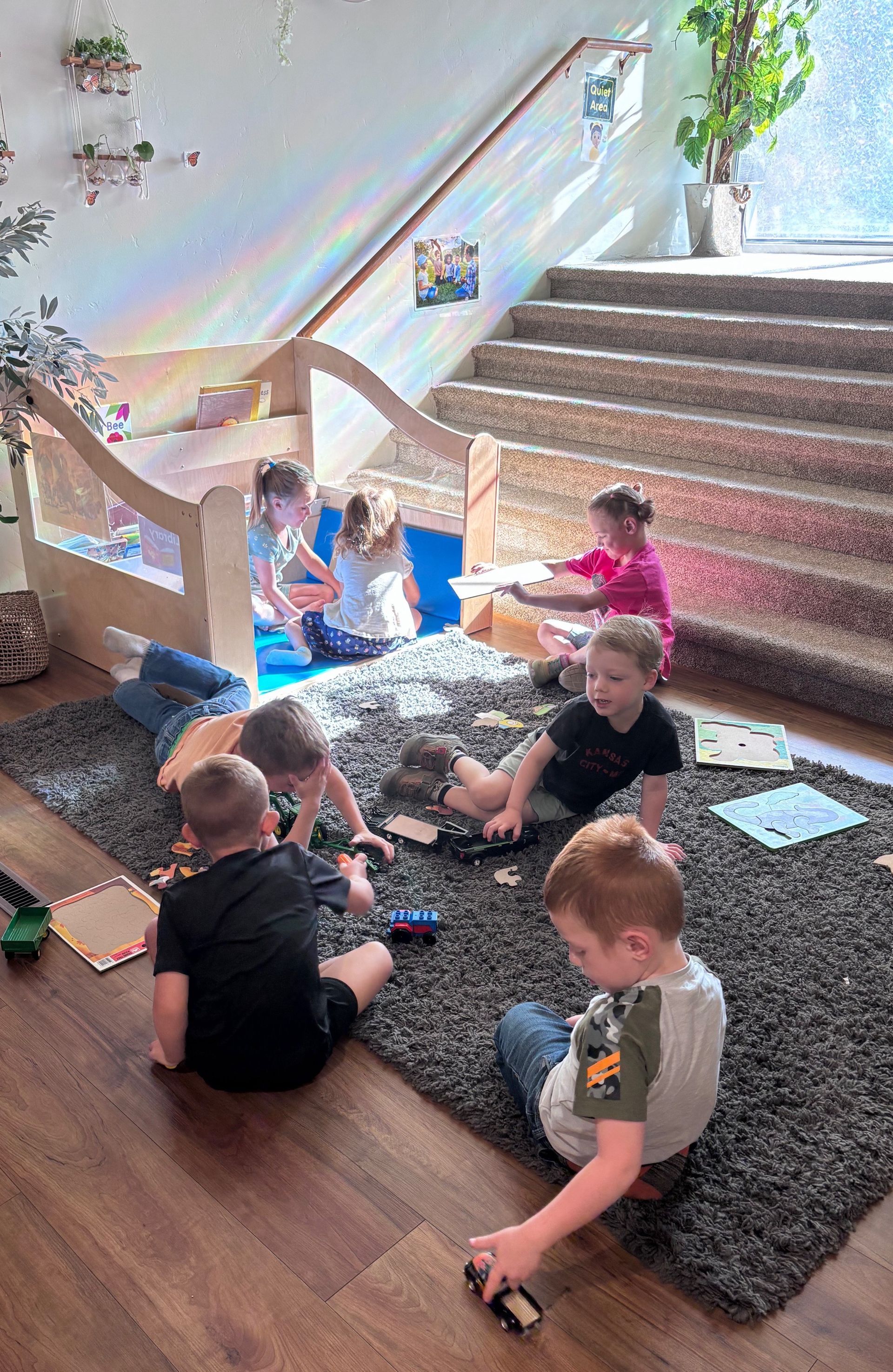 Children playing with toys on a rug; a wooden structure, stairs, and plants are in the background.