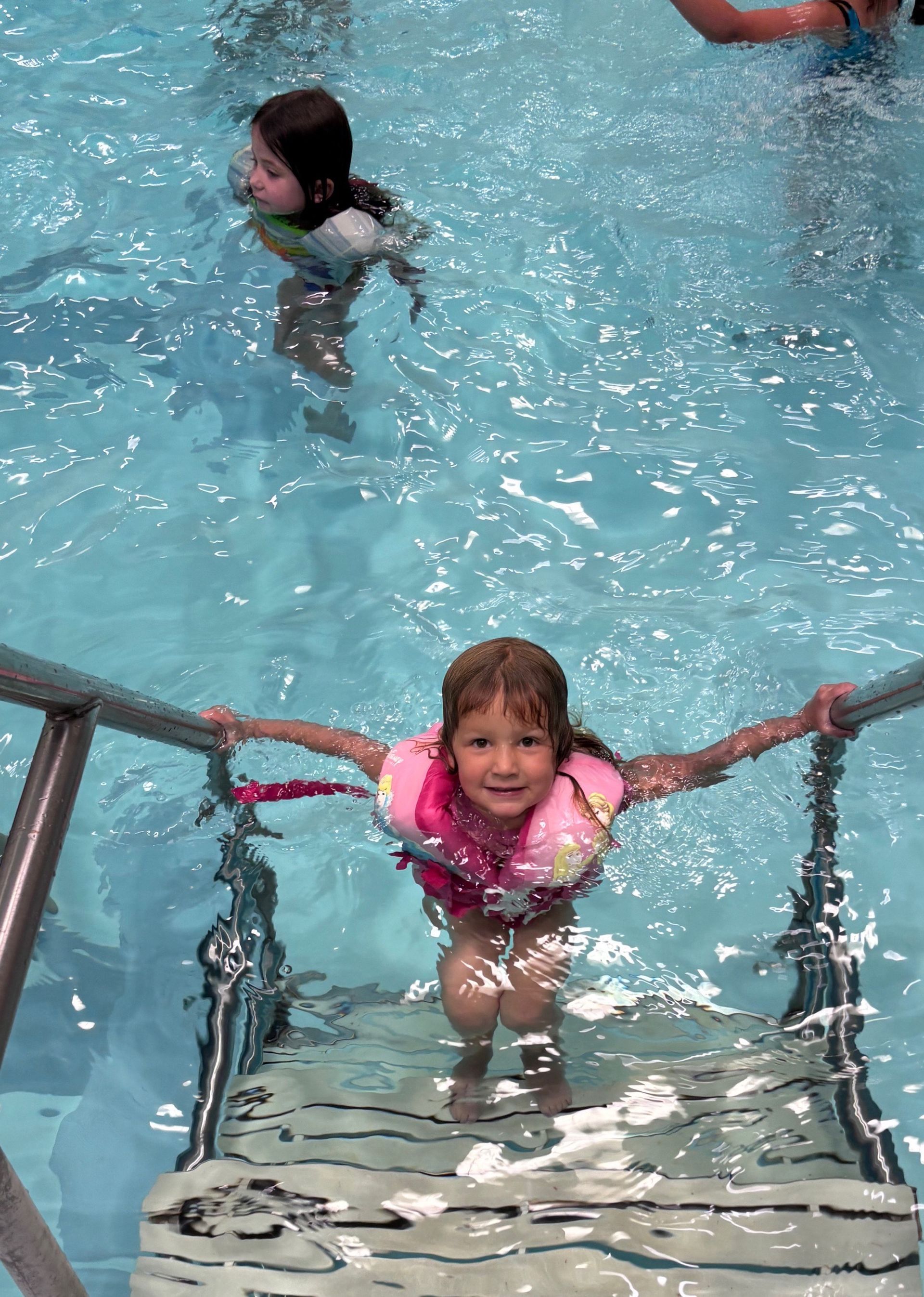 Child in a pink life jacket climbs out of a pool using metal steps, smiling. Another child swims in the background.