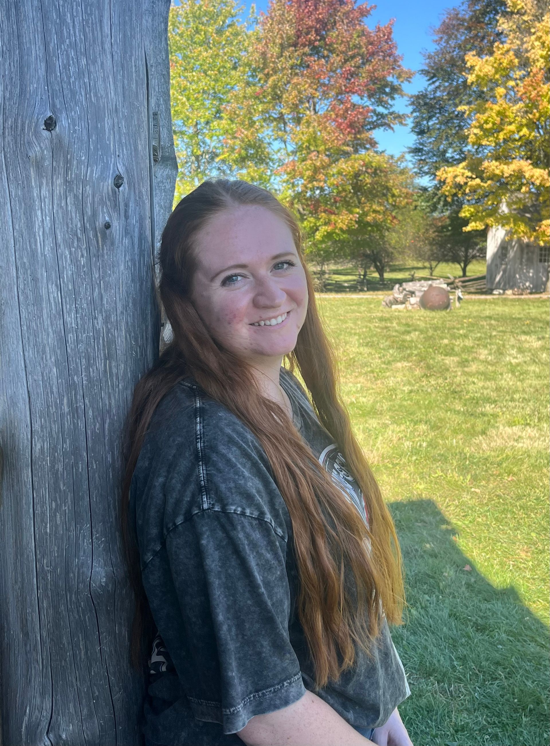 Woman with long red hair smiles, leans on weathered wooden wall outdoors; fall foliage in background.