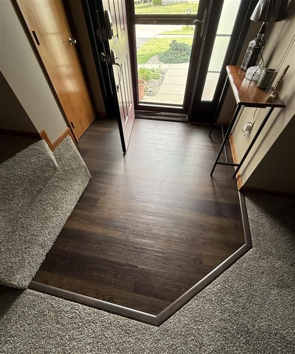 Indoor entryway with a wooden floor step down to gray carpet, leading to an open front door.