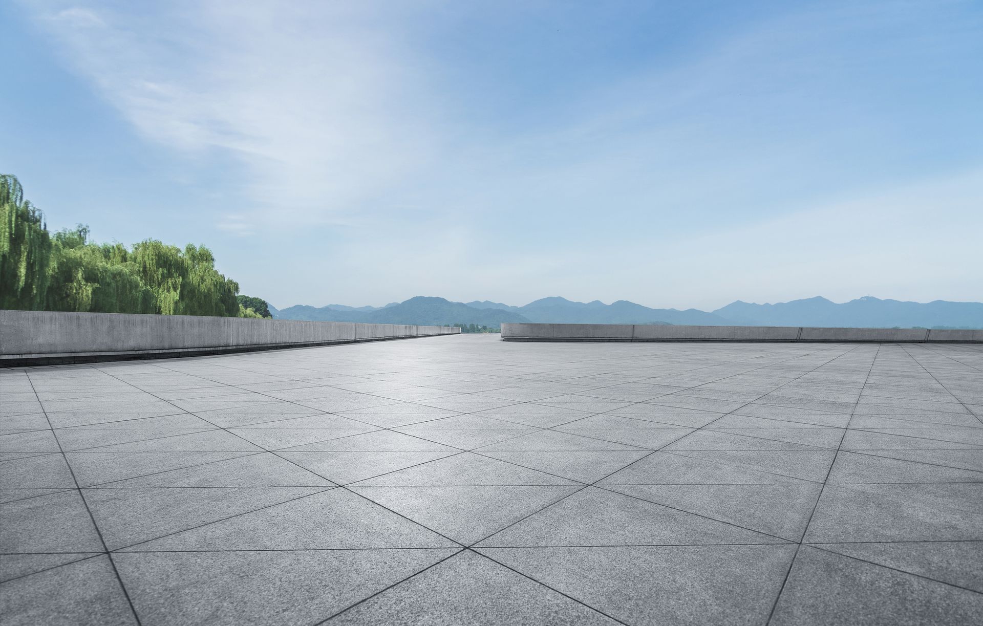 Empty concrete plaza with a view of distant mountains and a light blue sky. Trees line the left side.