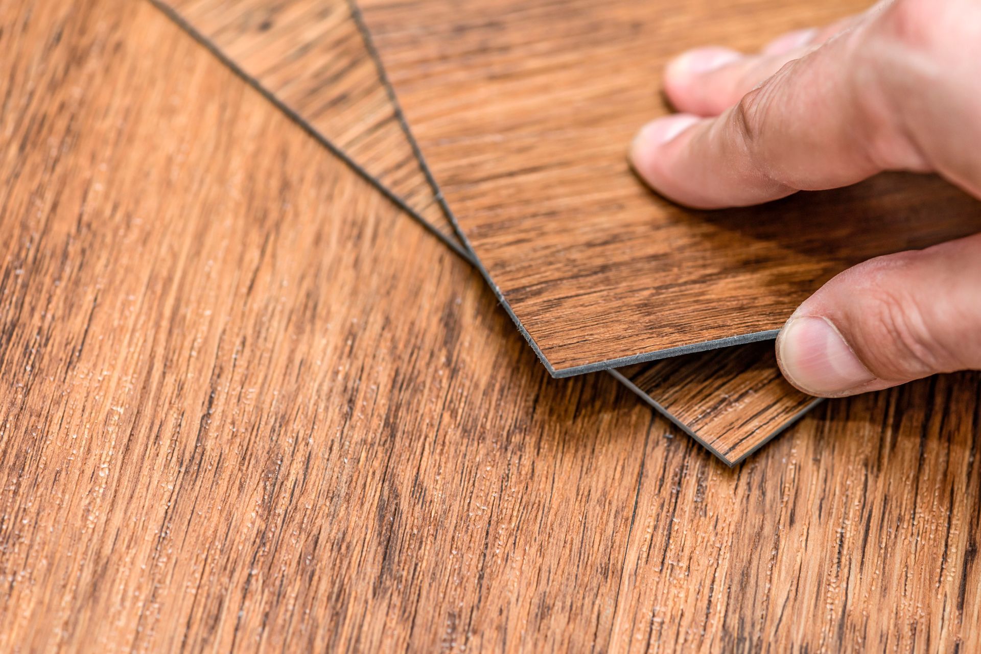 Hand holding up a sample of wood-look flooring, with a larger area of the same flooring visible.