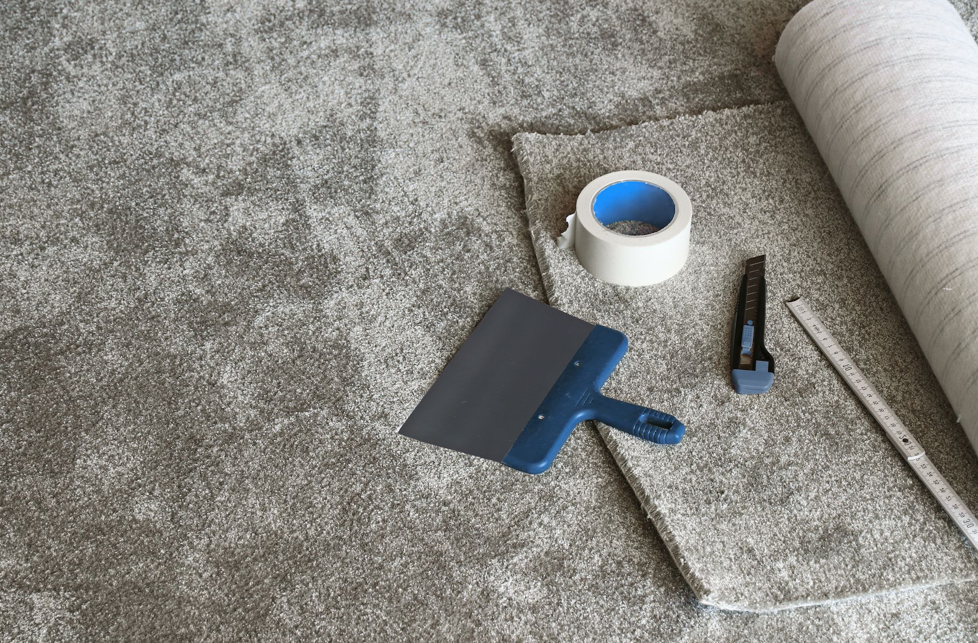 Carpet installation tools on a carpet, including a putty knife, utility knife, seam tape, and a roll of carpet.