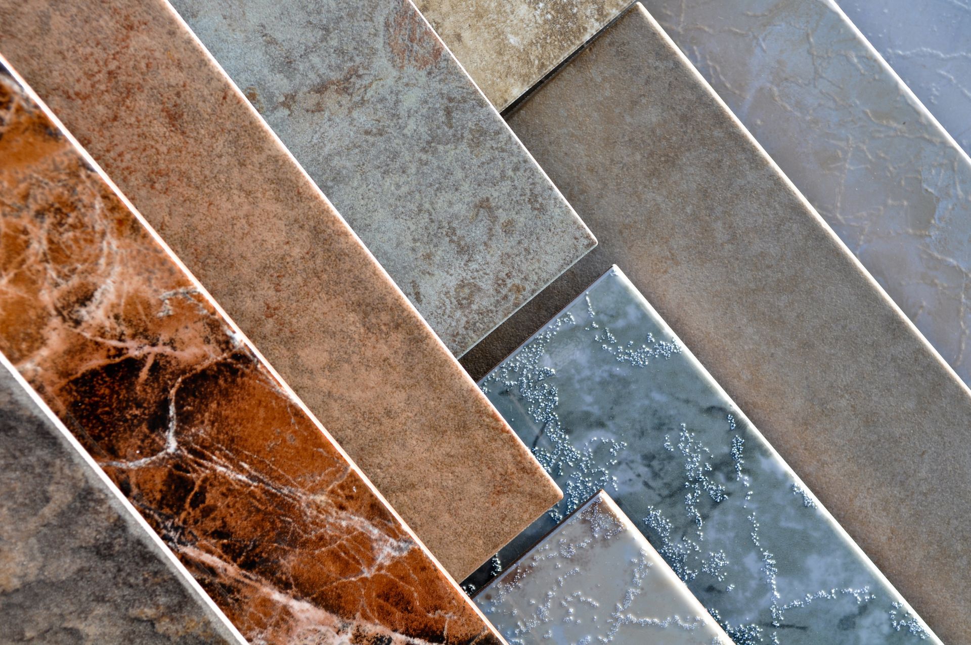 Close-up of a variety of rectangular tiles in shades of brown, gray, and beige arranged at an angle. The tiles showcase different textures and patterns.