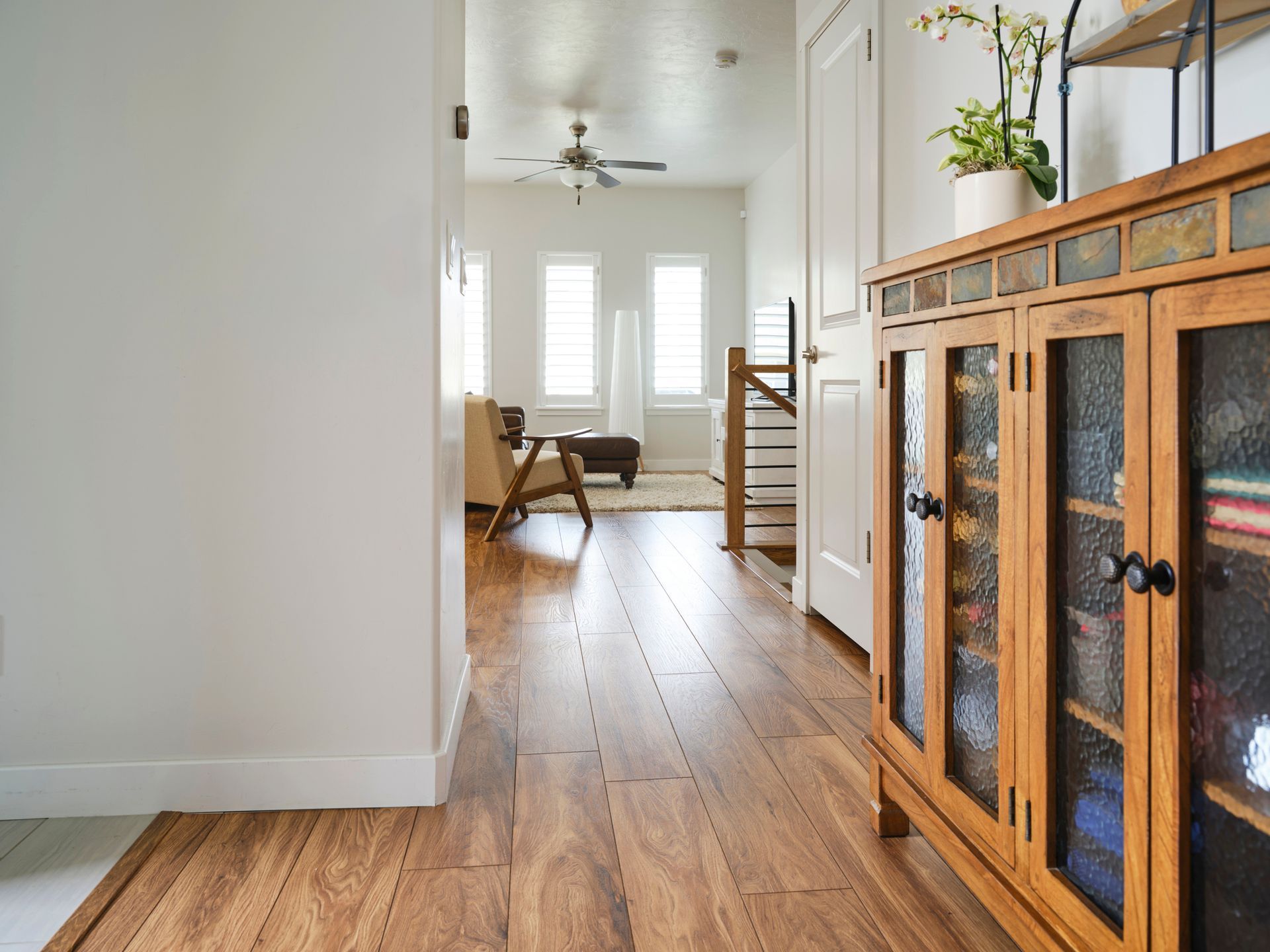 Wooden-floored hallway with an open view into a living area with furniture and natural light. A wooden cabinet sits in the foreground.