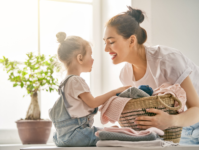 A person and a child smile at each other while folding clothes from a wicker basket near a potted plant.
