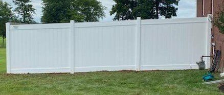 White vinyl fence in a grassy yard, trees in the background, near a brick building.