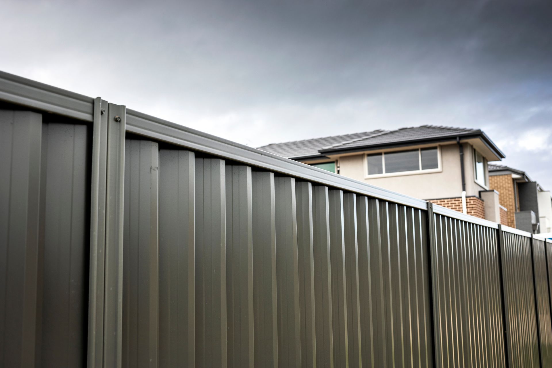 Modern metal fence with vertical panels in front of a brick house under cloudy skies.