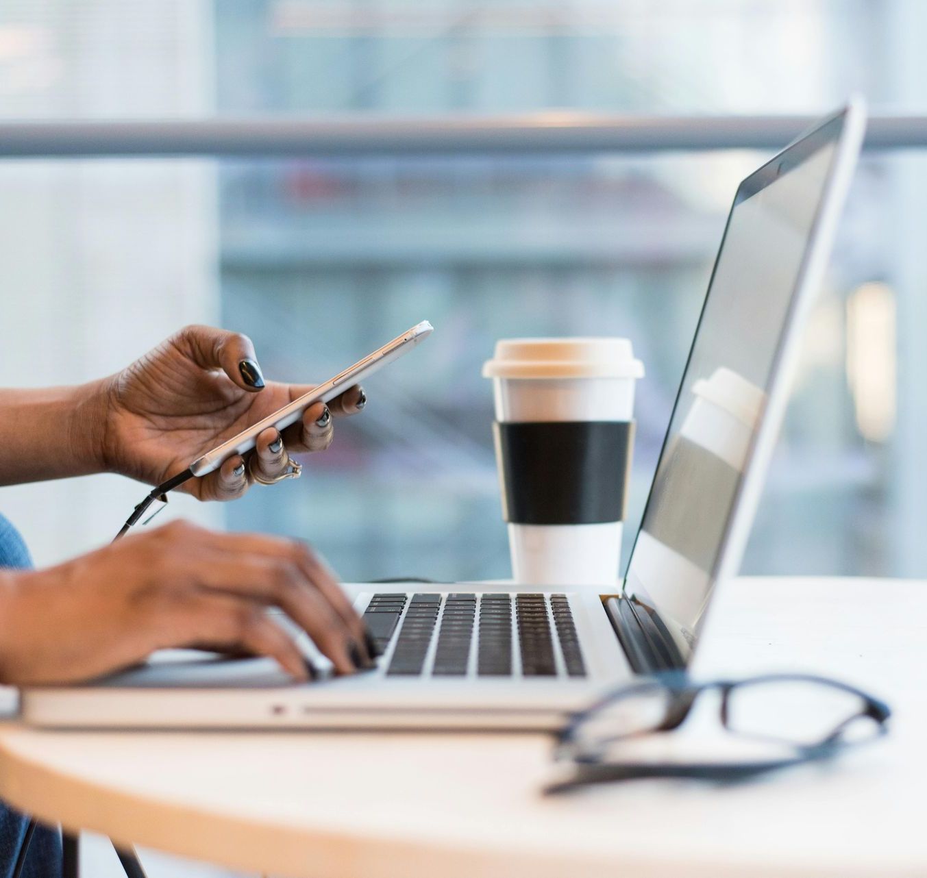 A person is typing on a laptop while holding a cell phone