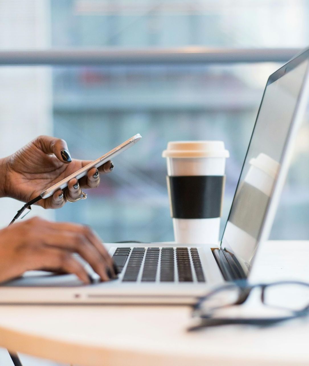 A person is typing on a laptop while holding a cell phone