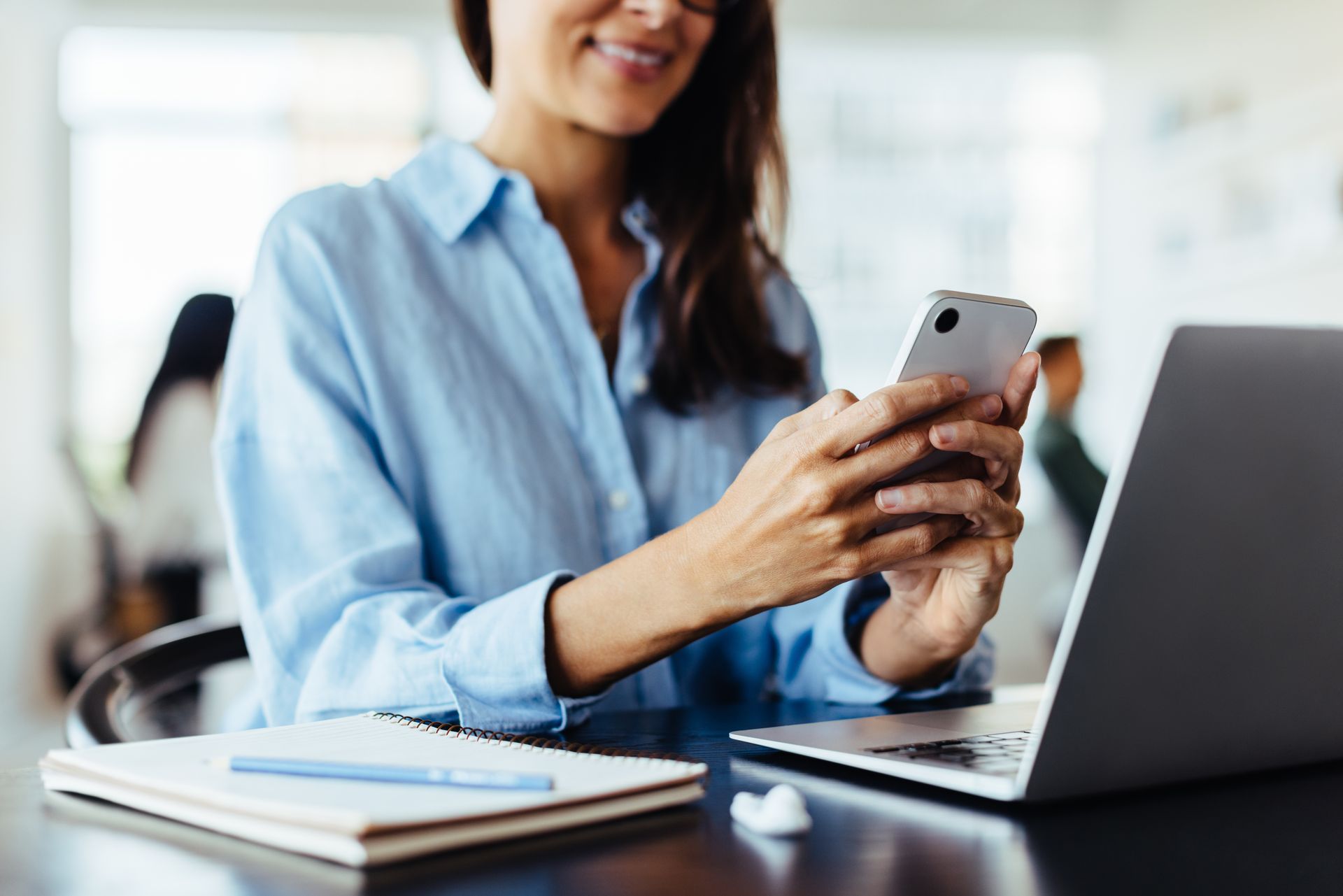 A woman is sitting at a desk using a laptop and a cell phone.